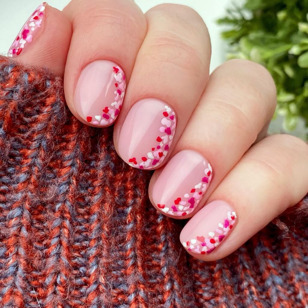 Short square blush pink nails with diagonal confetti hearts in red, pink and white at tips