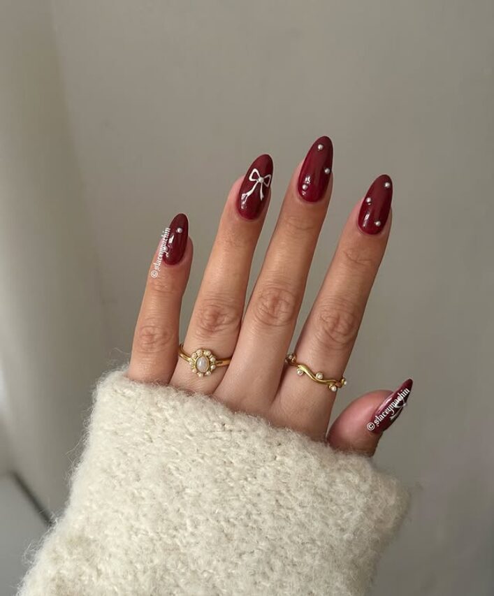 Hand with glossy burgundy nails featuring a white bow and small pearl accents