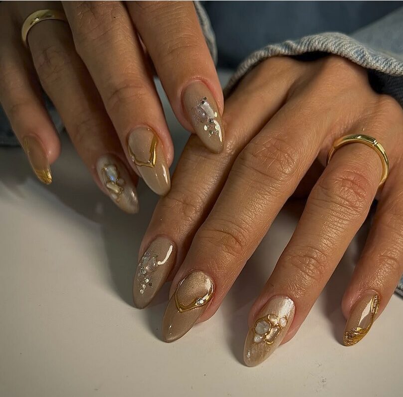Beige almond nails with gold foil accents and pearl-like embellishments