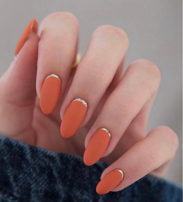 Hand with matte burnt-orange oval nails featuring a thin silver glitter cuticle