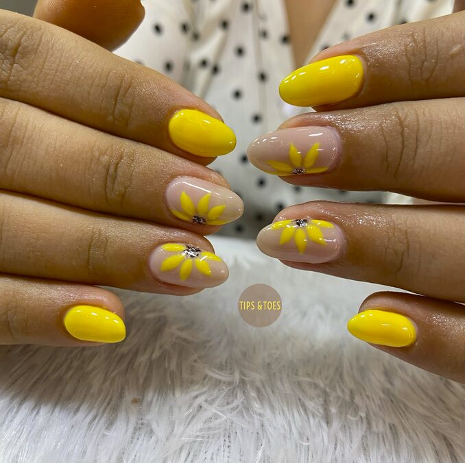 Glossy yellow nails with sunflower designs and rhinestone centers.
