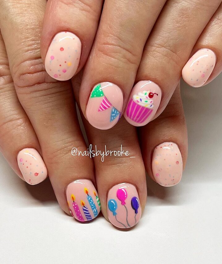 short peach-pink birthday nails featuring a cupcake, candles, balloons, party banners