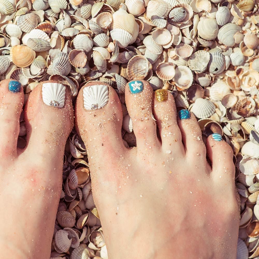 Beach-themed toe nails with 3D seashell art, starfish, gold glitter and blue glitter accents on sandy shore