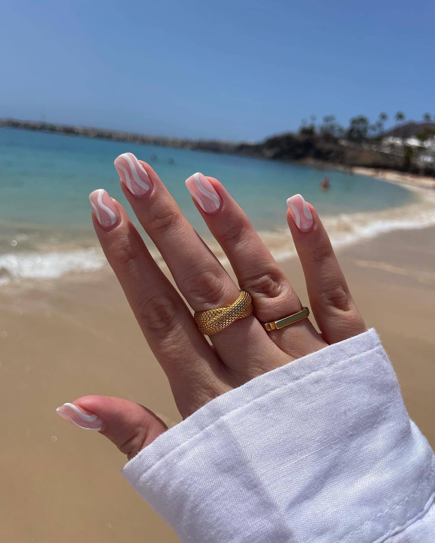 Sheer nude square nails with white wavy swirl lines at the beach