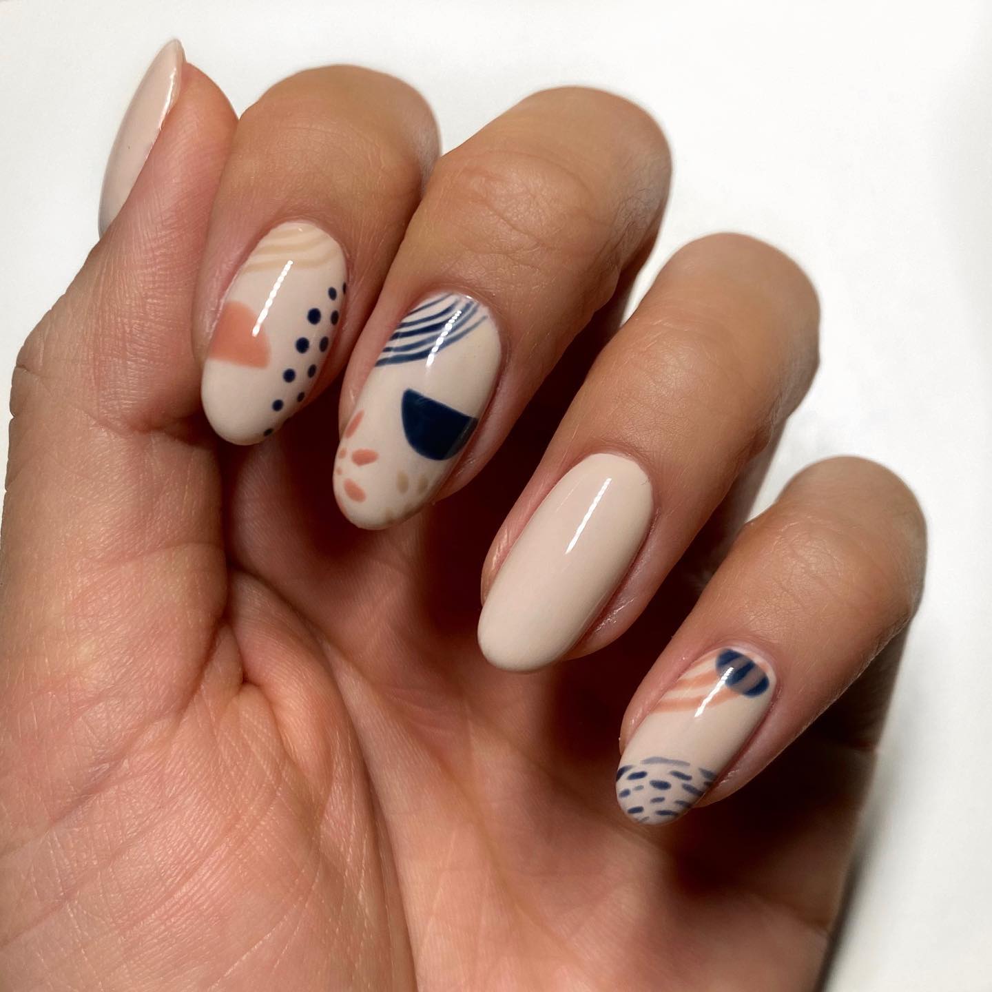 Short almond nails in creamy nude with abstract painted shapes — navy blue stripes, terracotta brushstrokes, dark dots, and geometric half-circles