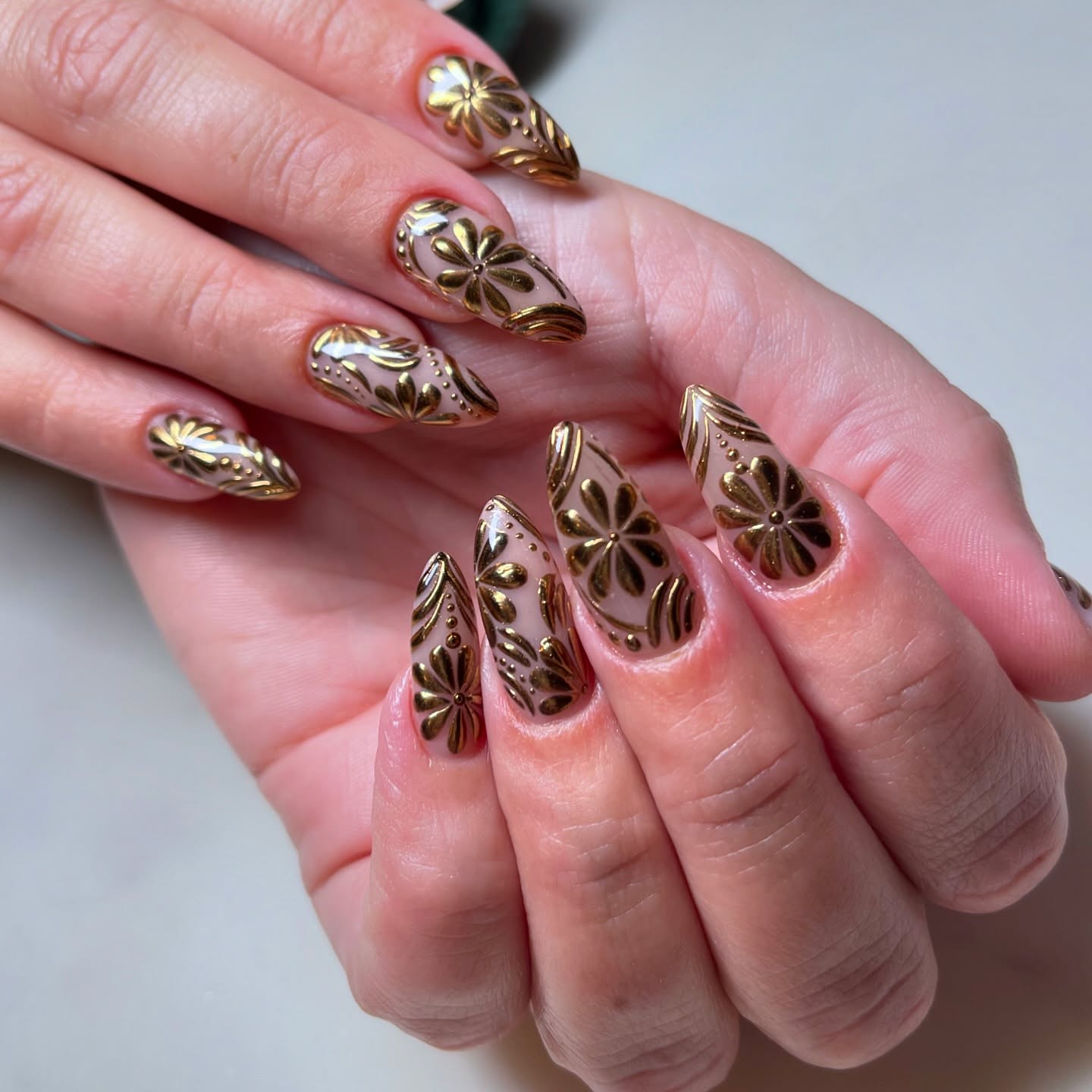 Long almond nails in warm nude with raised antique gold 3D floral and leaf relief designs covering the full nail surface