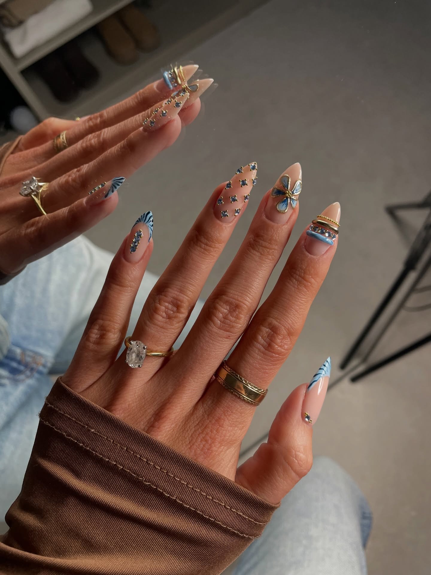 Long almond nails with blue swirls, scattered rhinestones, and a gold butterfly charm on a nude base