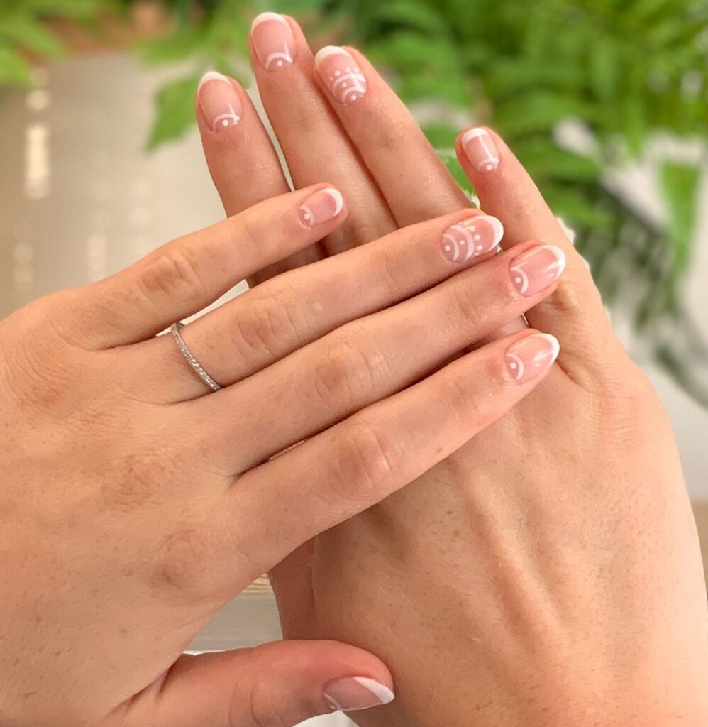 Short oval nails with natural sheer base, clean white French tips, and white dotwork crescent and mandala accent detail