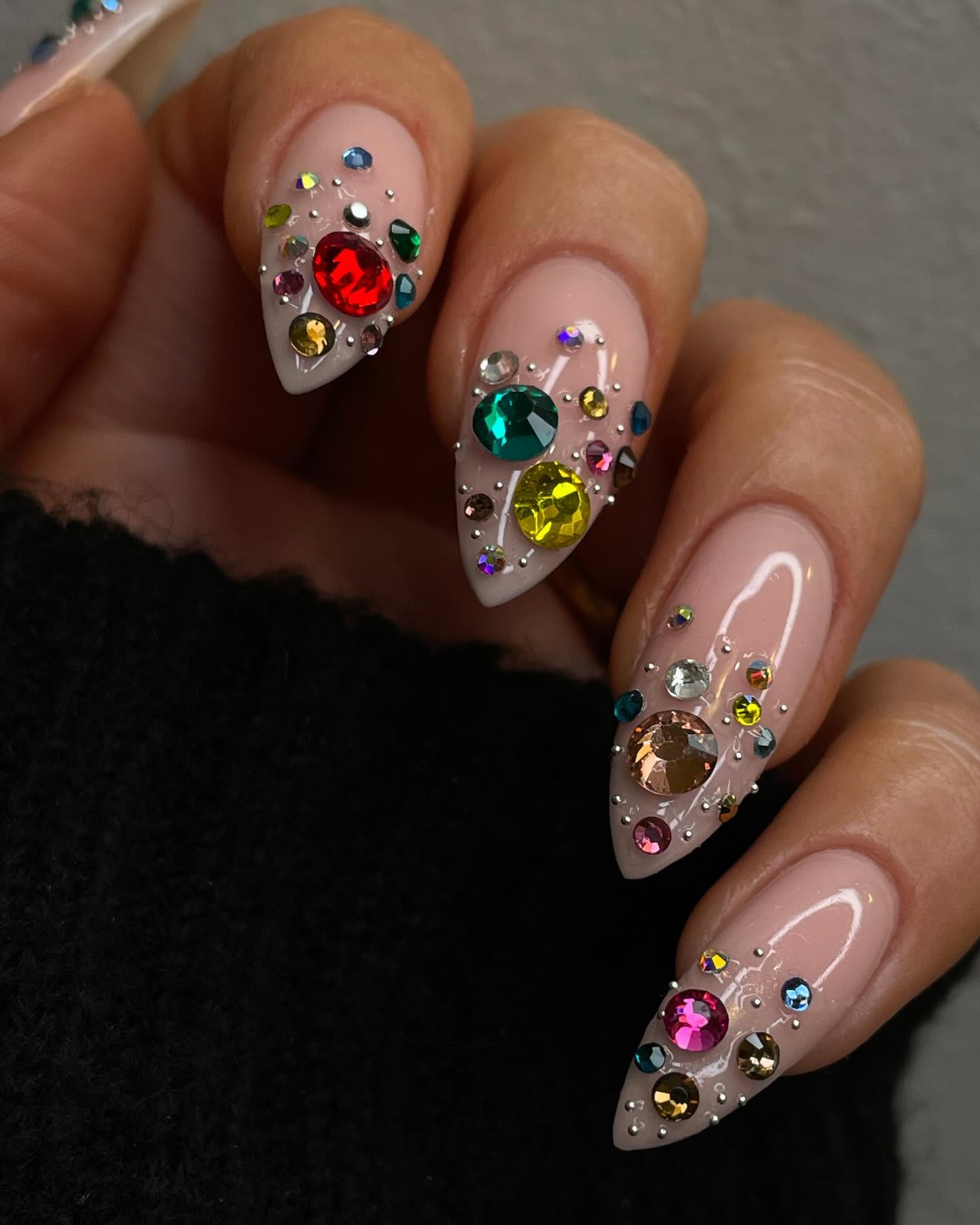 Sheer nude almond nails loaded with oversized multicolour rhinestones in a scattered pattern