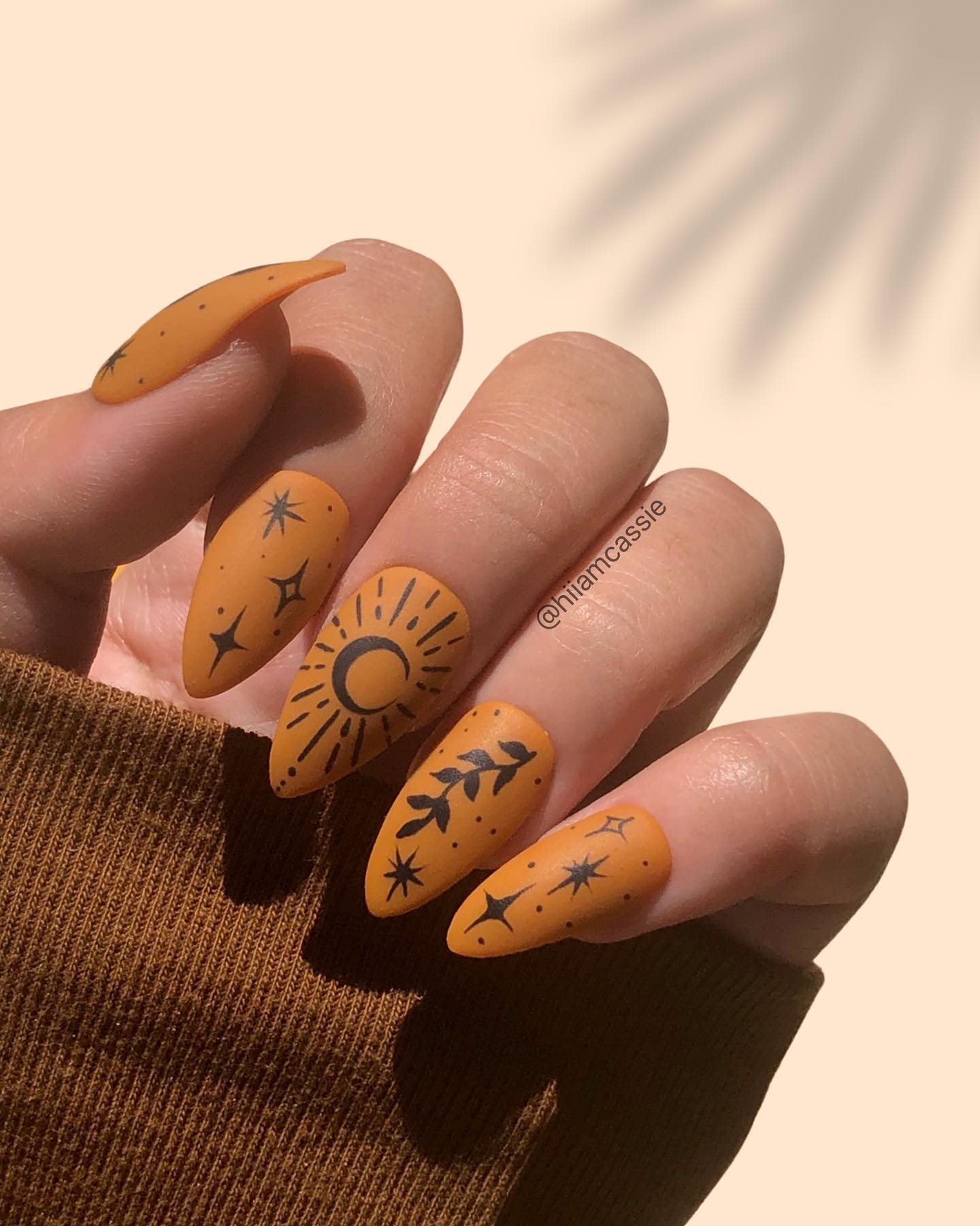 Long almond nails in matte burnt orange with black hand-painted sun, crescent moon, star, and botanical motifs