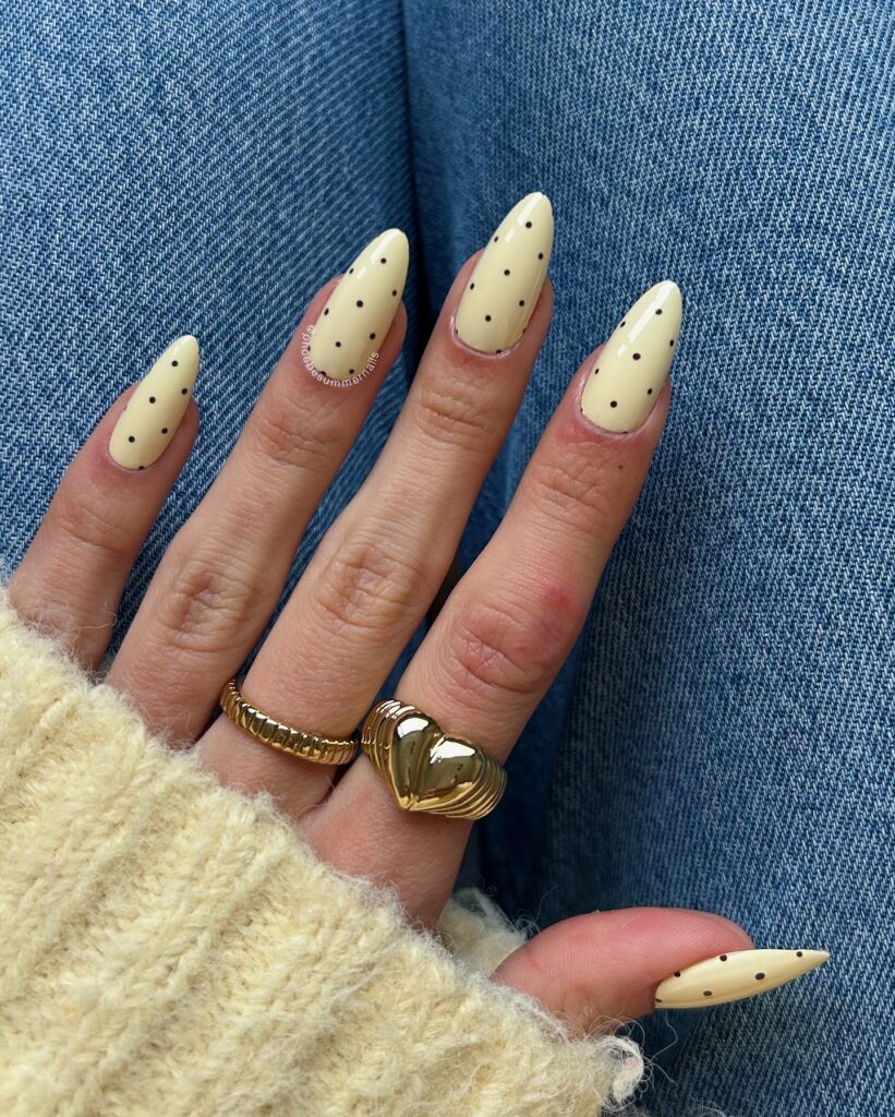 Long almond nails in soft butter yellow scattered with evenly spaced tiny black polka dots in a vintage fabric-inspired design