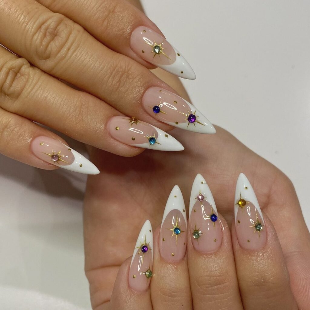 Long stiletto nails with white French tips, scattered gold star charms, and multicoloured gemstone rhinestones on a nude base