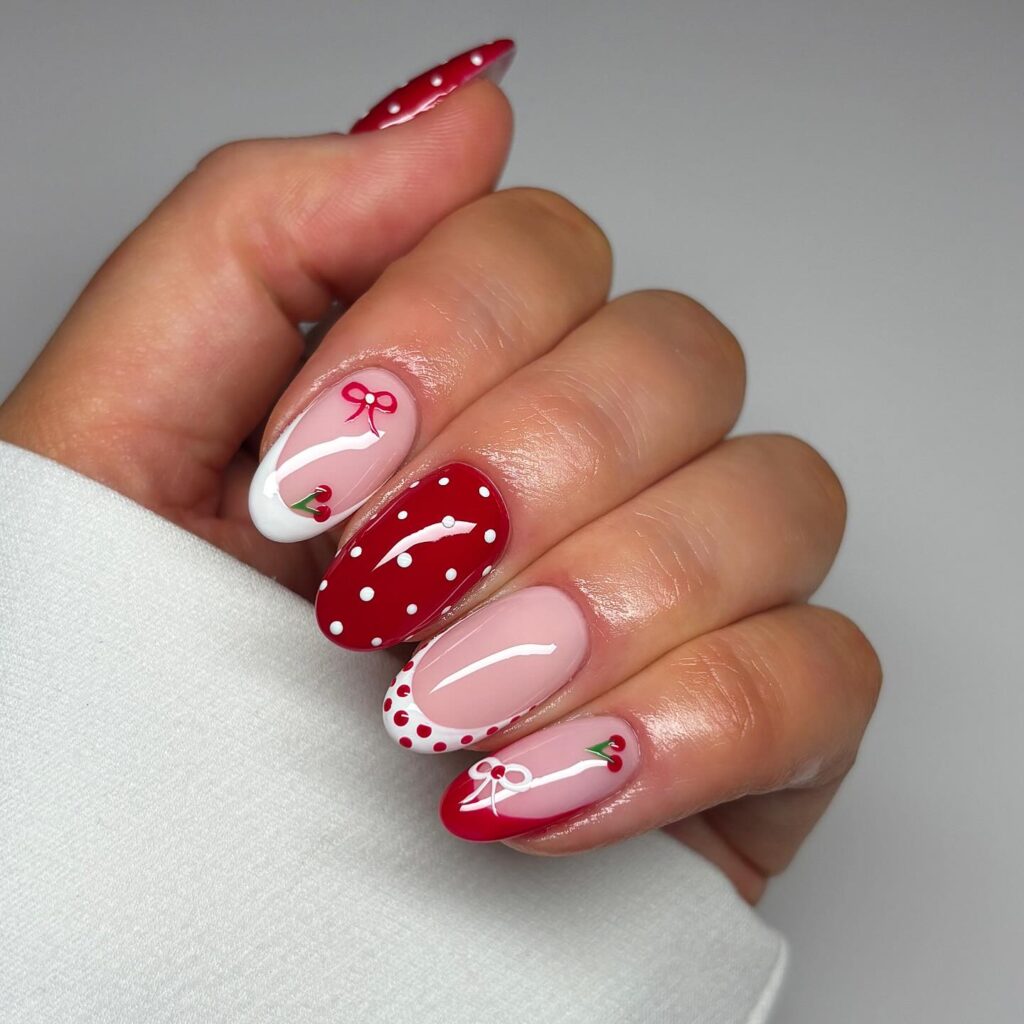 Oval nails mixing solid red, sheer pink with red polka dots, and delicate cherry and bow hand-painted accent details