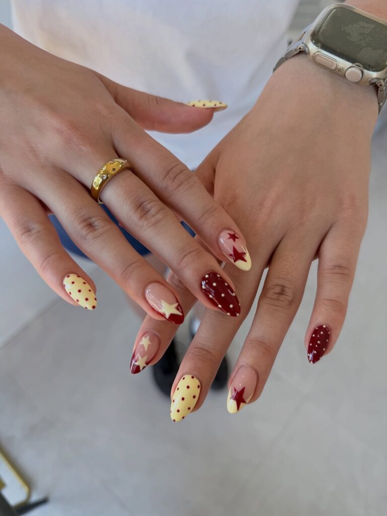 Almond nails in cream and deep red featuring gold polka dots and red star accent details in a retro diner-inspired style