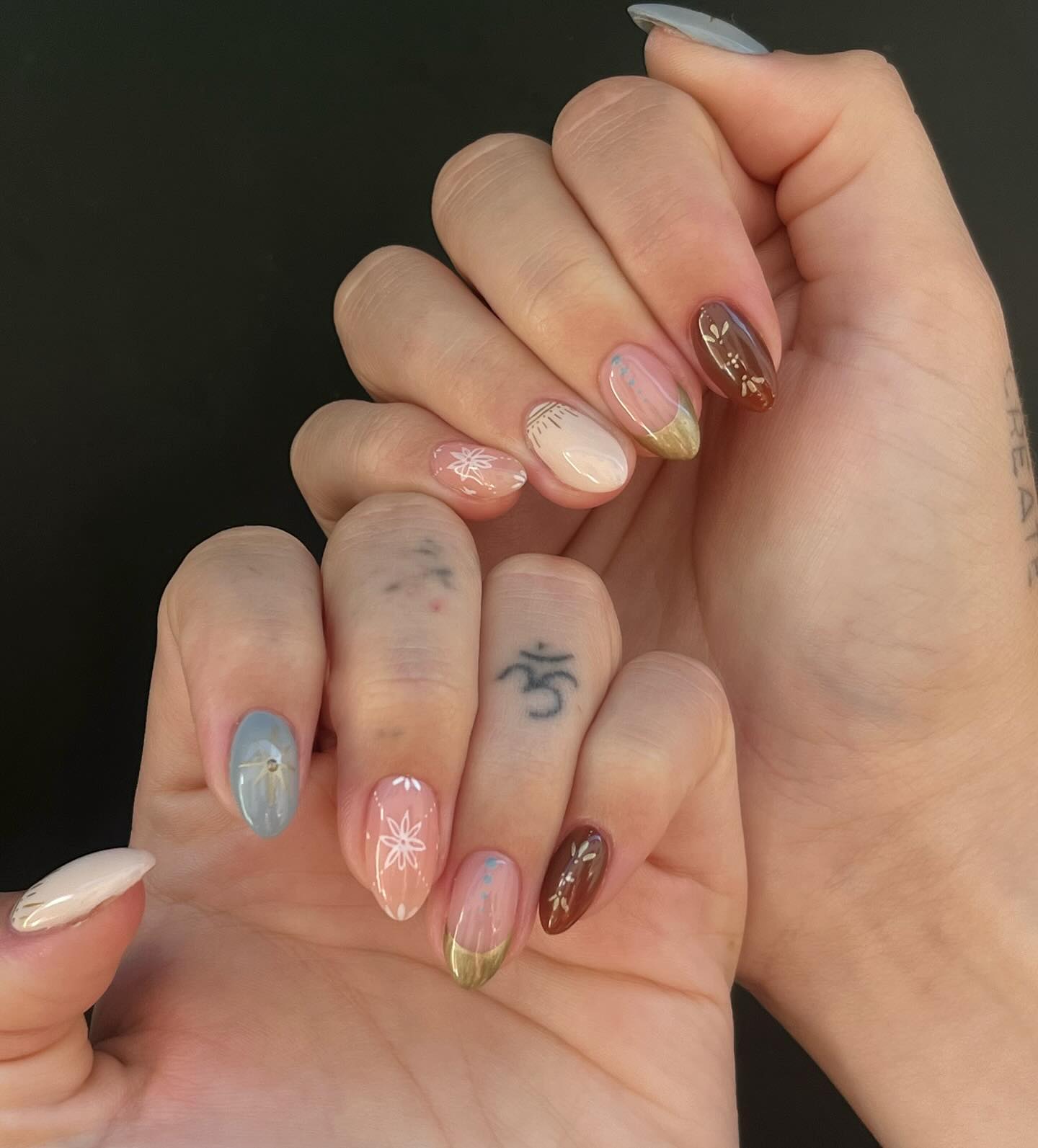 Almond nails in mixed earthy tones — slate blue, blush pink, terracotta brown — with delicate white floral motifs and gold foil tips
