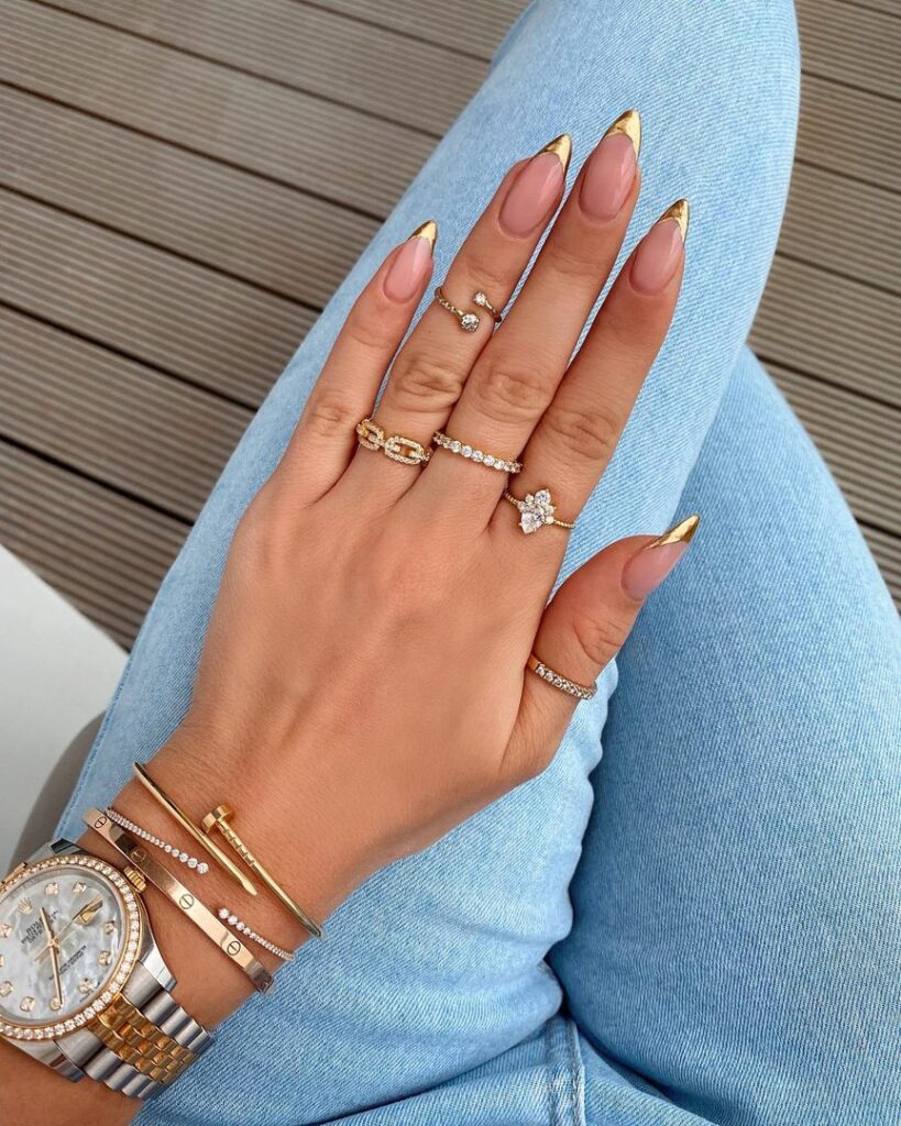 Long oval nails with gold chrome mirror tips on a nude base, styled with gold jewellery