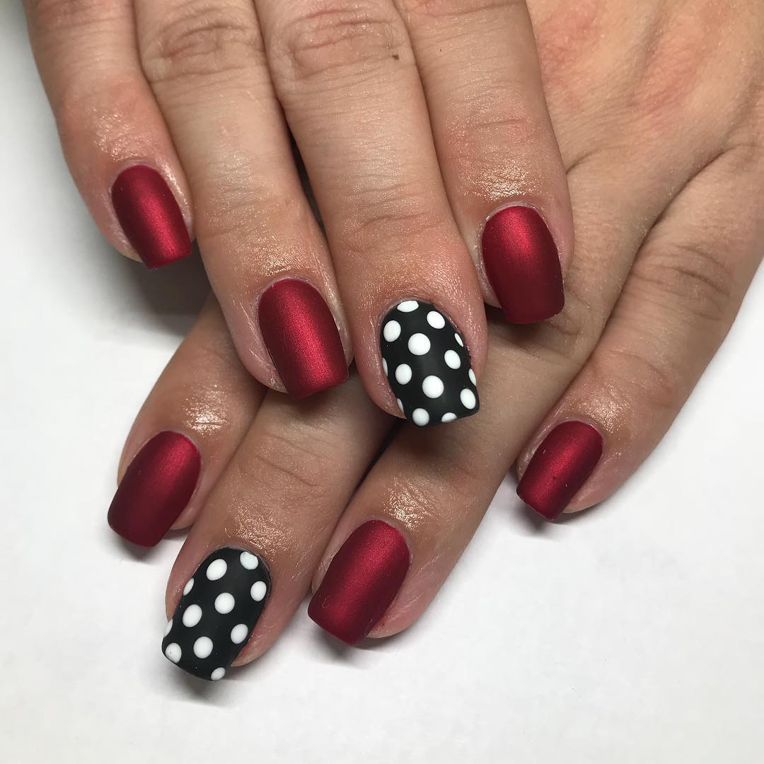 Short round matte dark red nails with black accent nails covered in white polka dots