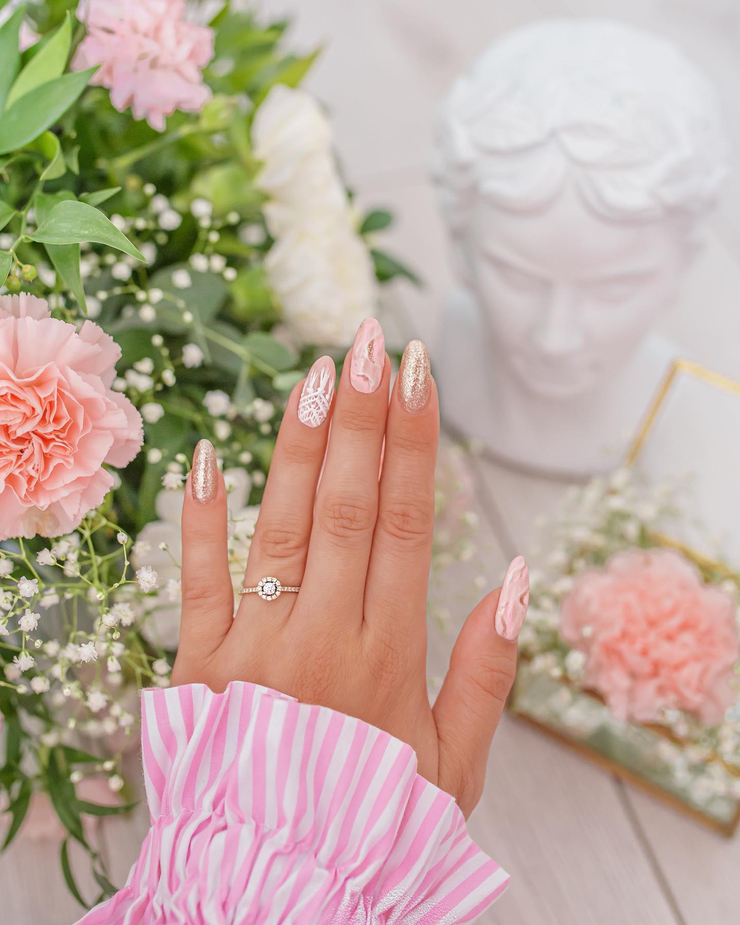 Long almond nails combining pearlescent pink, gold glitter, and white lace detail against a floral backdrop
