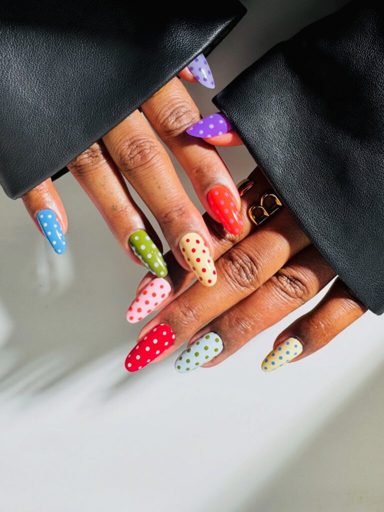 Long almond nails each in a different bold base colour — purple, red, green, cream, and blue — covered in contrasting white polka dots