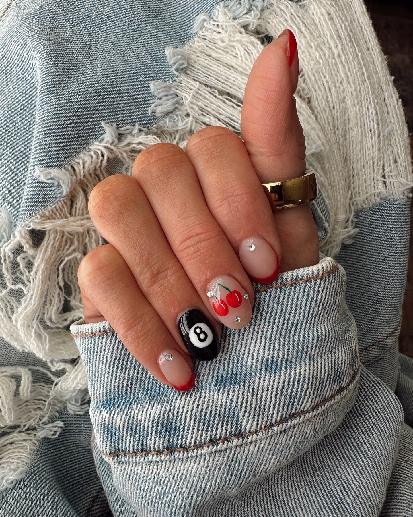 Short round nails with red tips, a black 8-ball accent nail, and red cherry nail art with rhinestone details