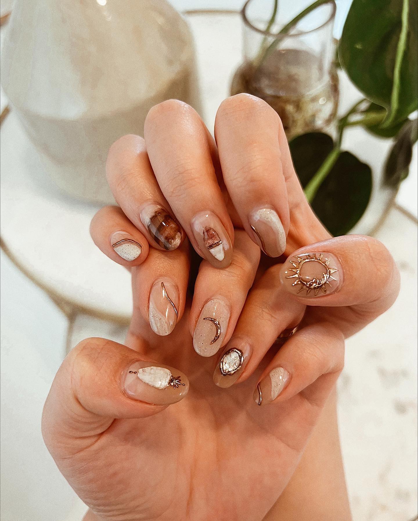 Short oval nails in sandy beige with embedded crystal fragments, gold crescent moon charm, gold sun charm, and wire-like line details