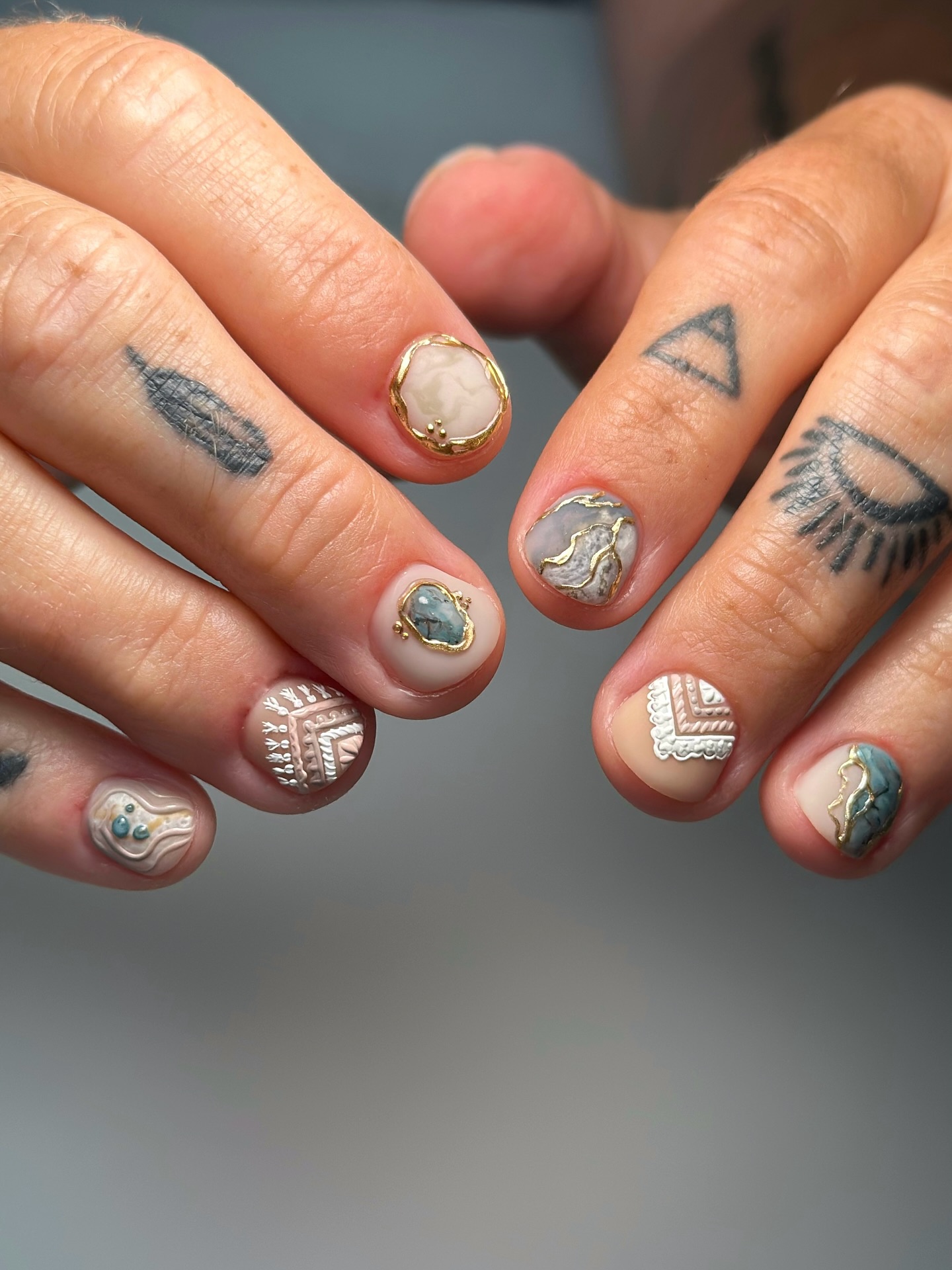 Short round nails with embedded turquoise and marble stone cabochons framed in gold, aztec tribal white patterns on nude matte base
