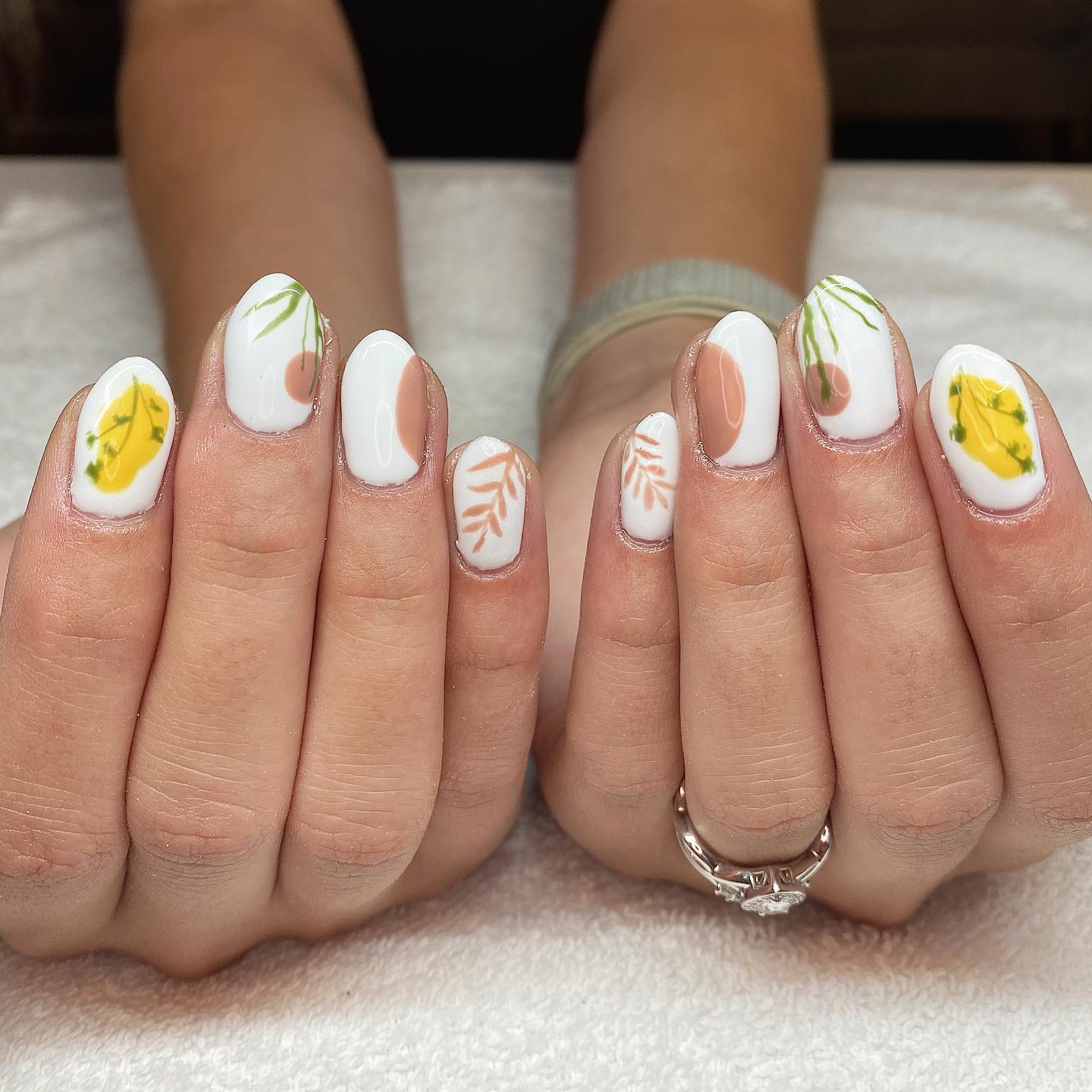 Short oval nails in white with terracotta moon phase shapes, hand-painted green botanical sprigs, and yellow floral accents