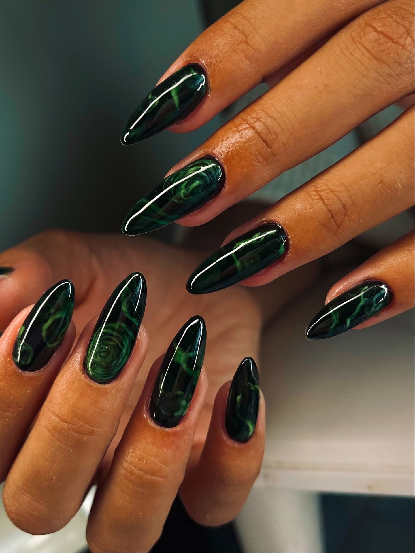 Long almond nails are finished in a deep green-and-black glossy design with subtle reflective detailing