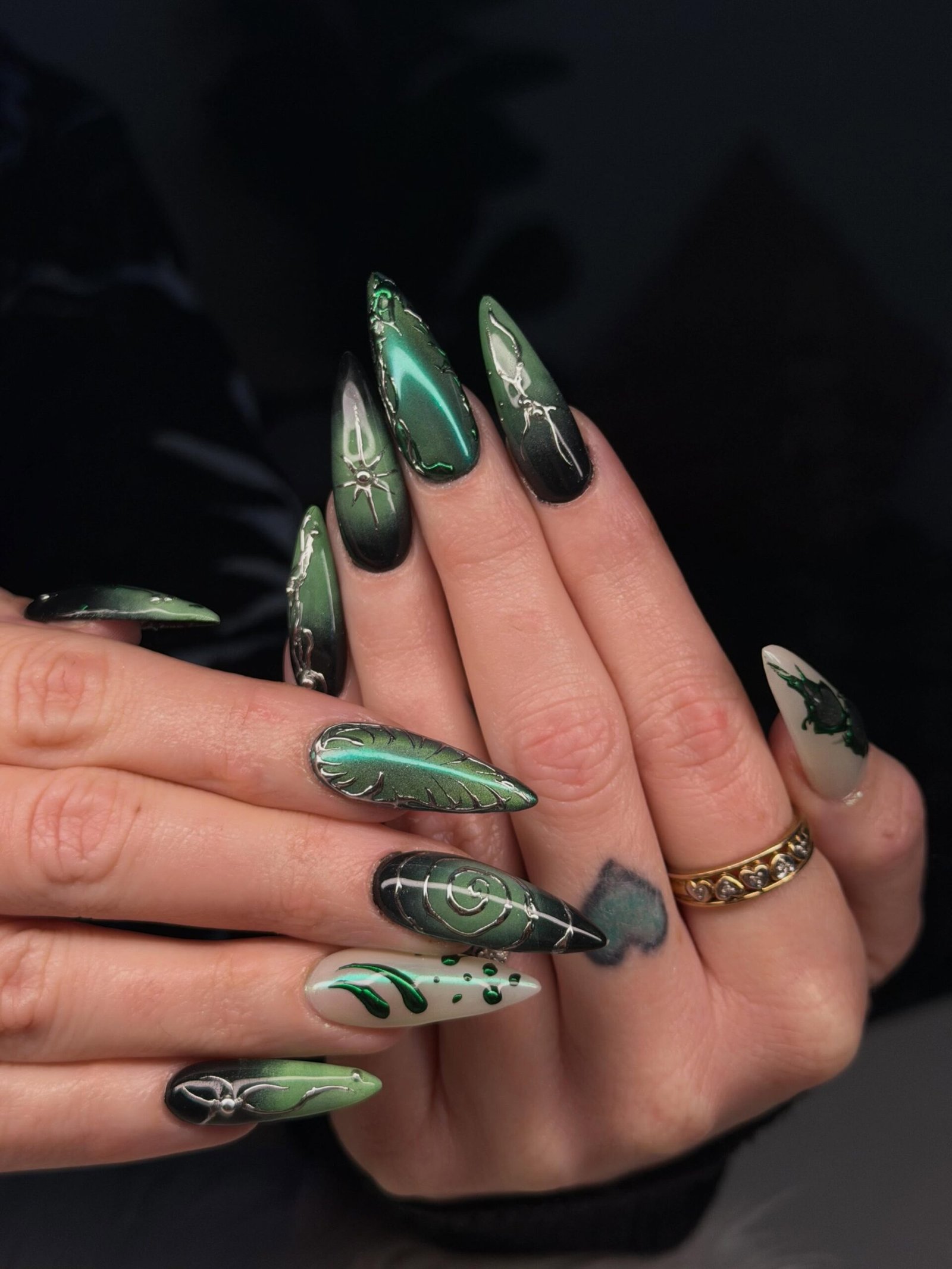 Long stiletto nails use deep green and black tones with contrasting patterned and metallic-looking details