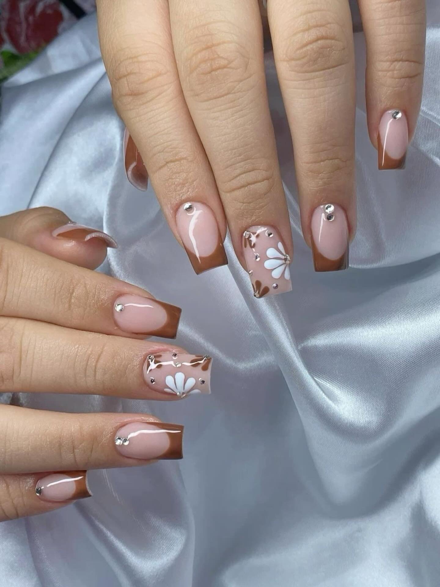 Medium-length pink-toned nails feature chocolate French tips alongside delicate white floral details and small metallic accents.