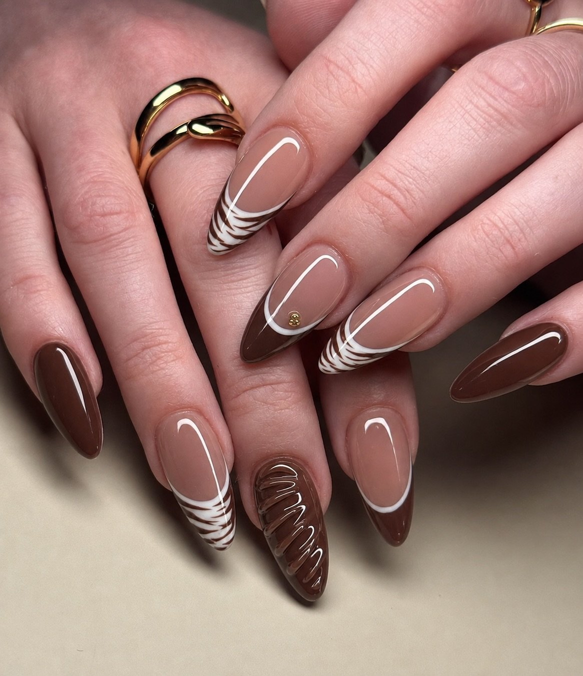 Long almond nails combine chocolate-brown and nude tones with thin double white French curves and patterned brown accents.