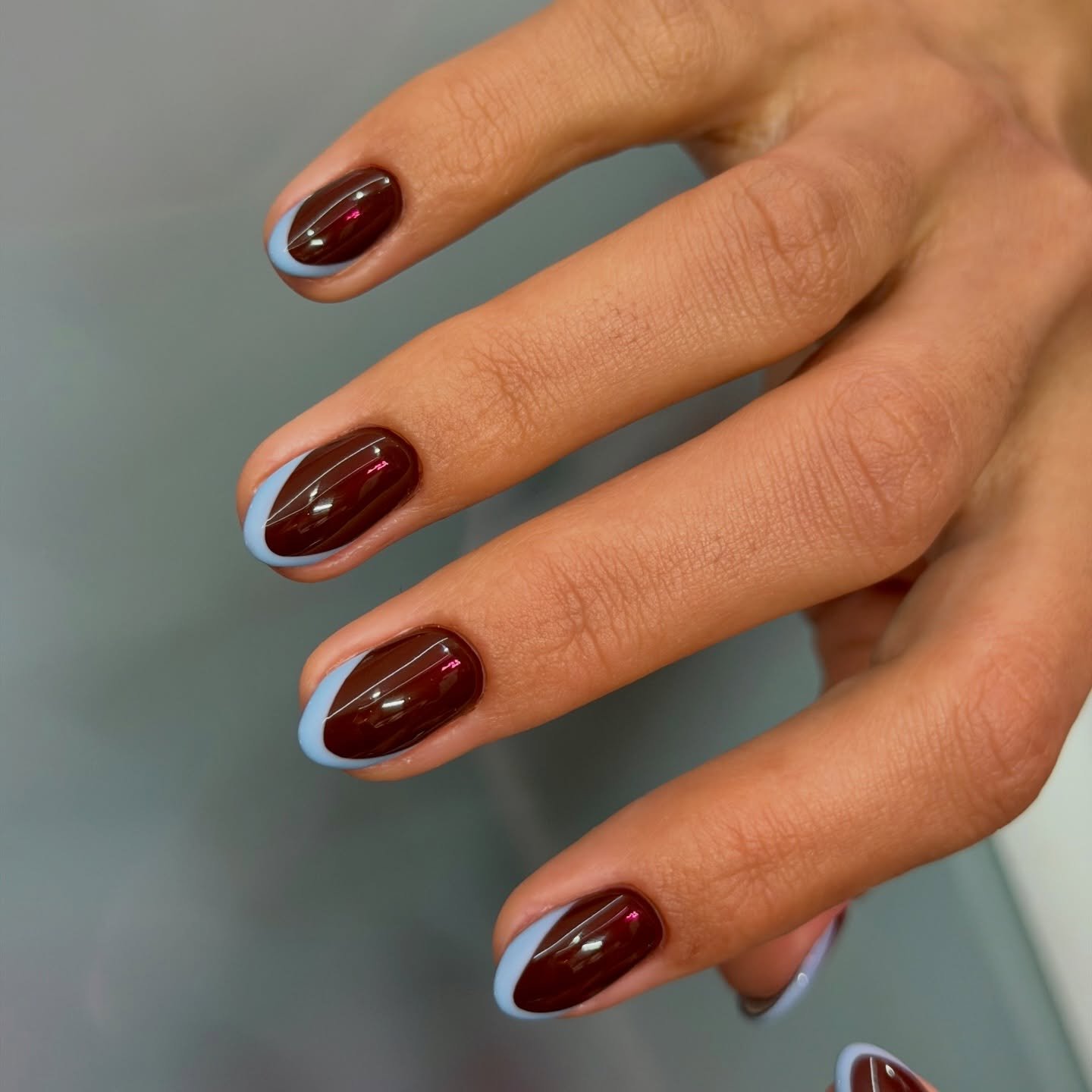 Short rounded nails are painted deep chocolate brown and finished with a slim pale blue-white French edge.