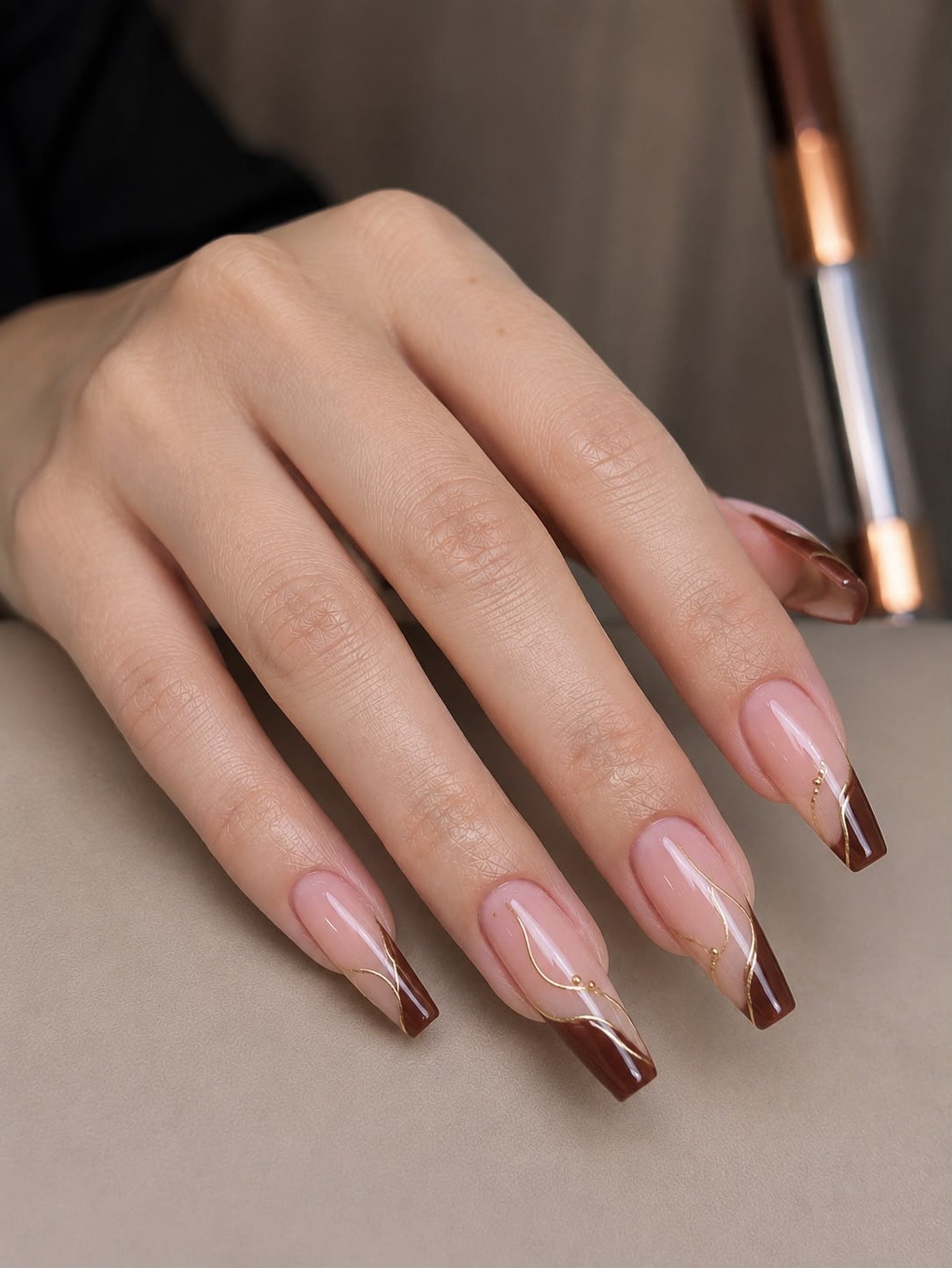 Long almond nails use a sheer pink-nude base with glossy chocolate-brown French tips and fine accent detailing.