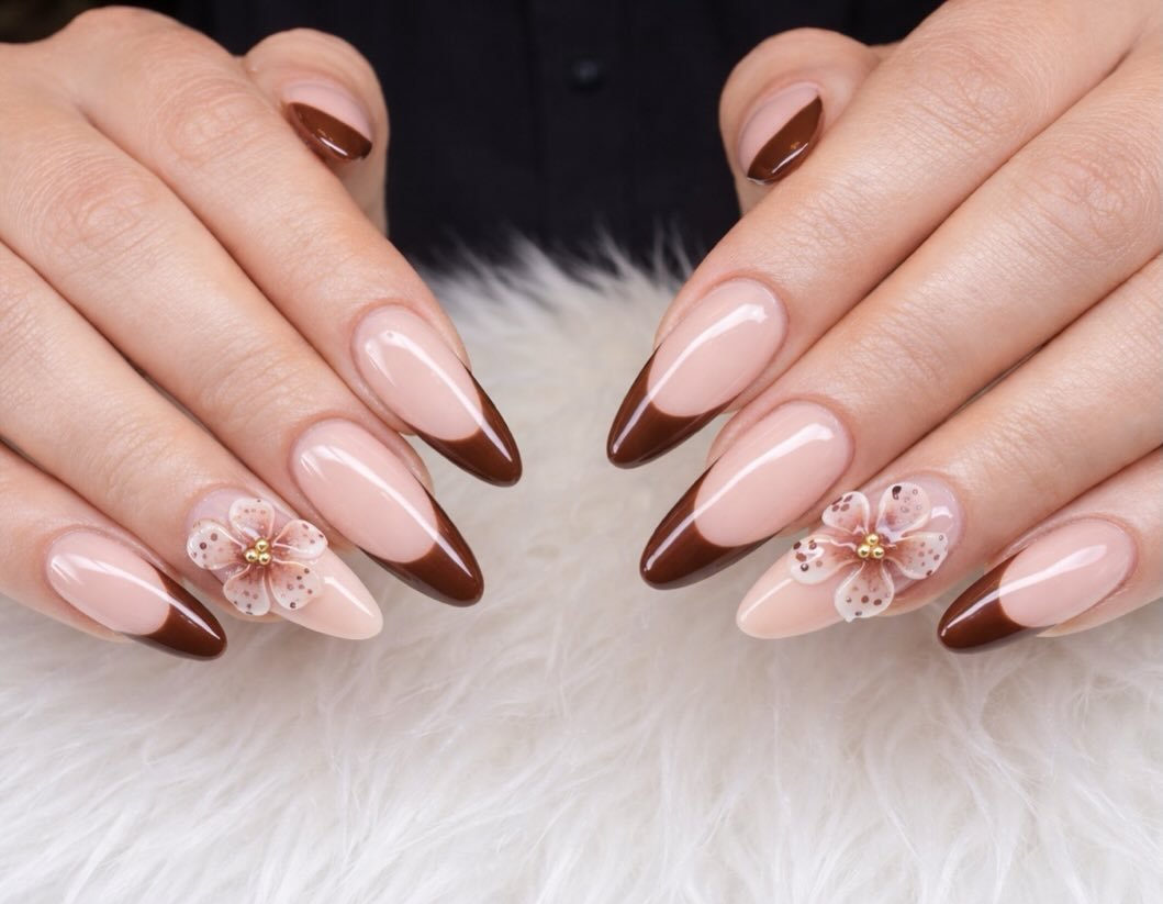 Long almond nails feature pale blush bases, chocolate-brown French tips, and delicate pale floral accents.