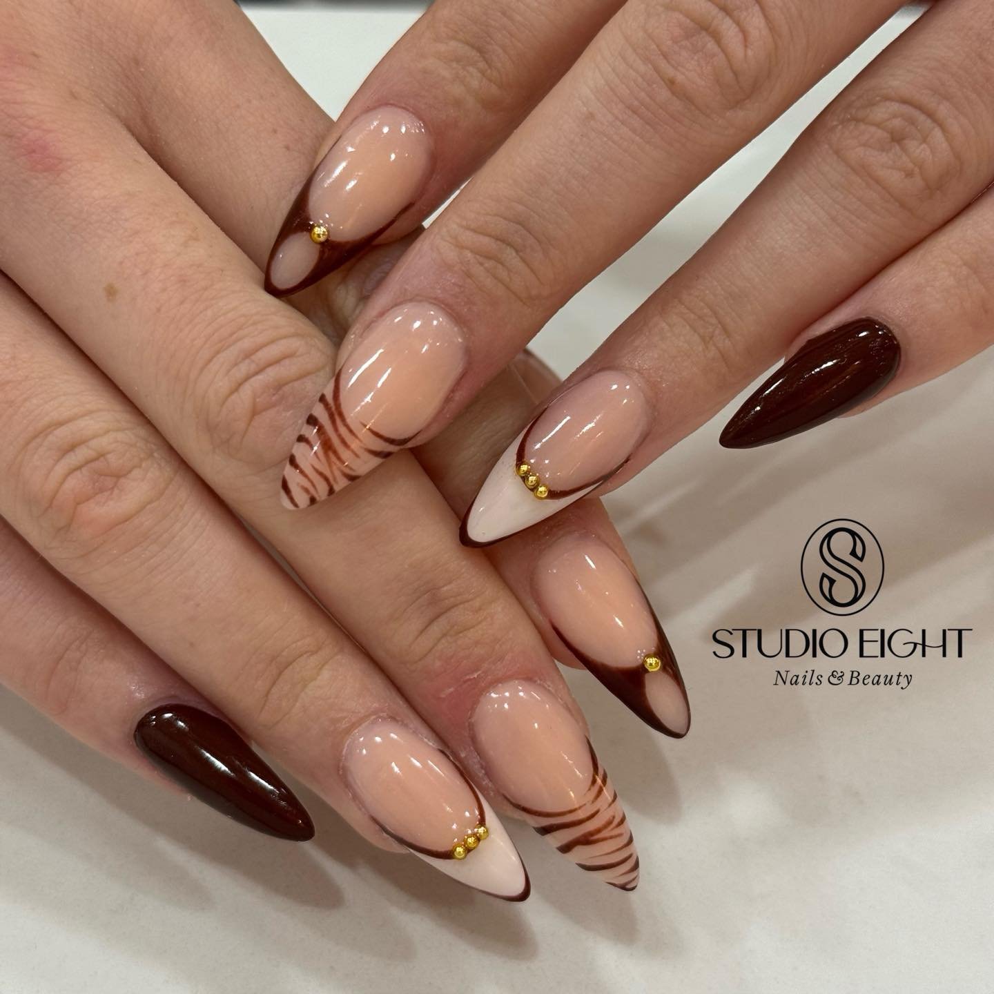 Medium almond nails pair a nude base with chocolate-brown French tips and small gold-toned decorative details.