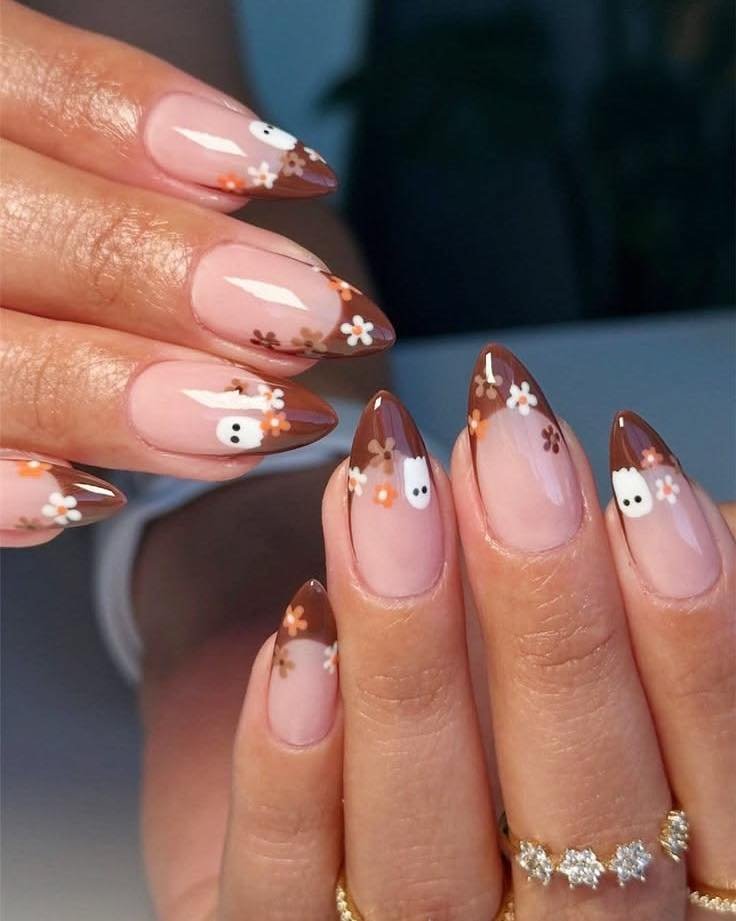 Long almond nails use a pale pink base with chocolate-brown French tips, tiny white floral motifs, and gold-toned dots.