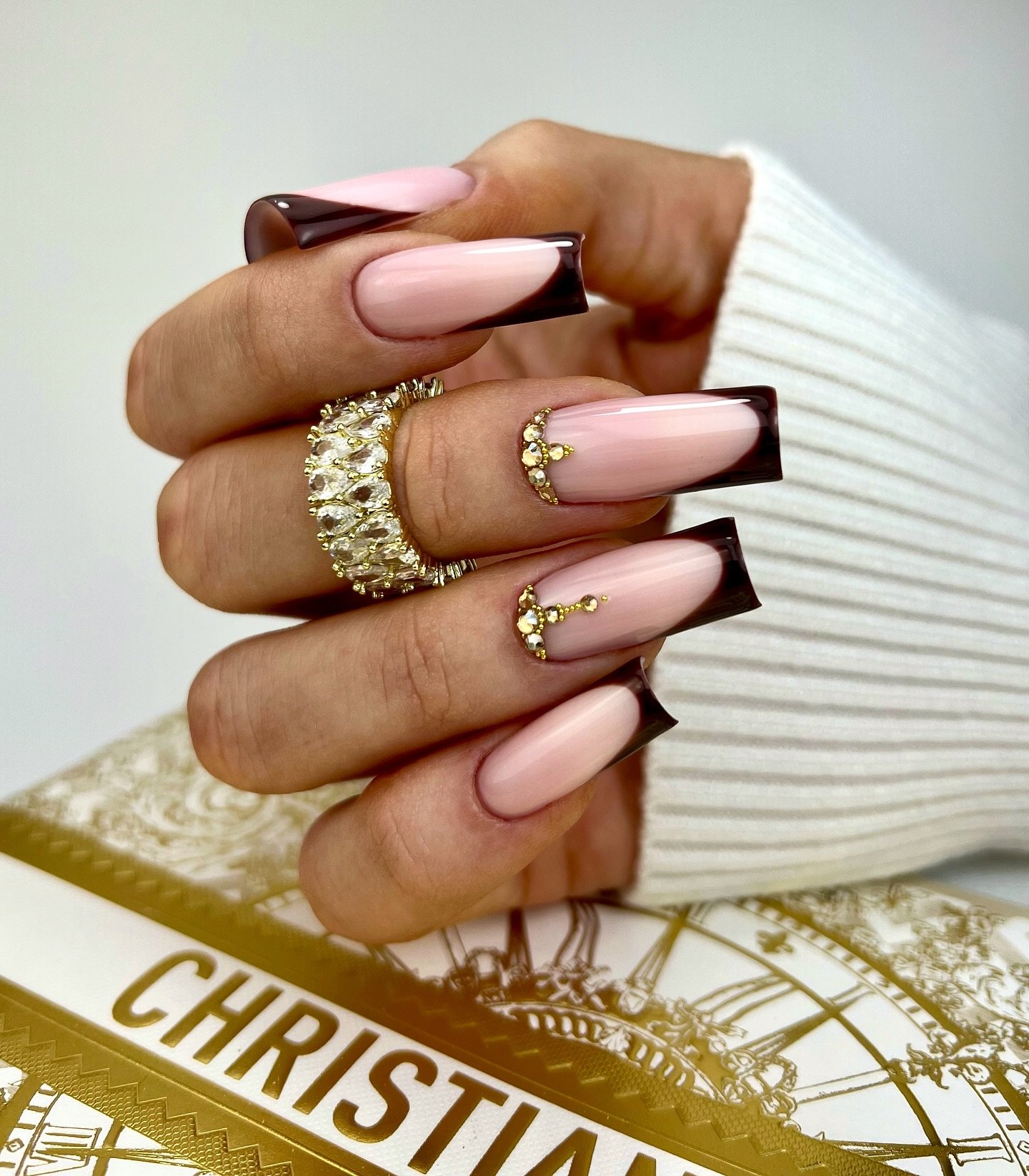 Long square nails combine a pale pink base, dark chocolate French tips, and small gold-toned embellishments.
