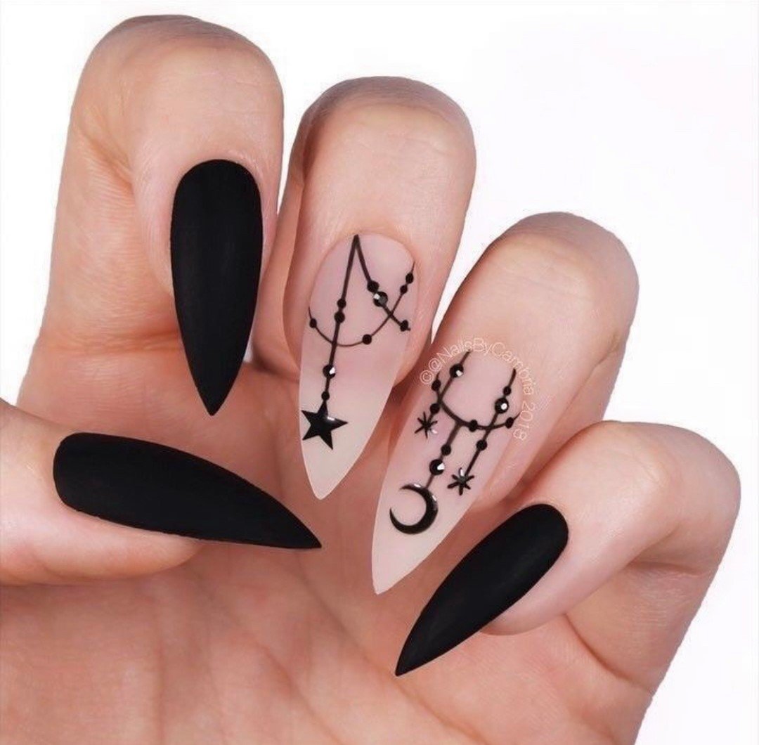 Long nude nails feature matte black pointed sections alongside thin black occult-style symbols and stars.