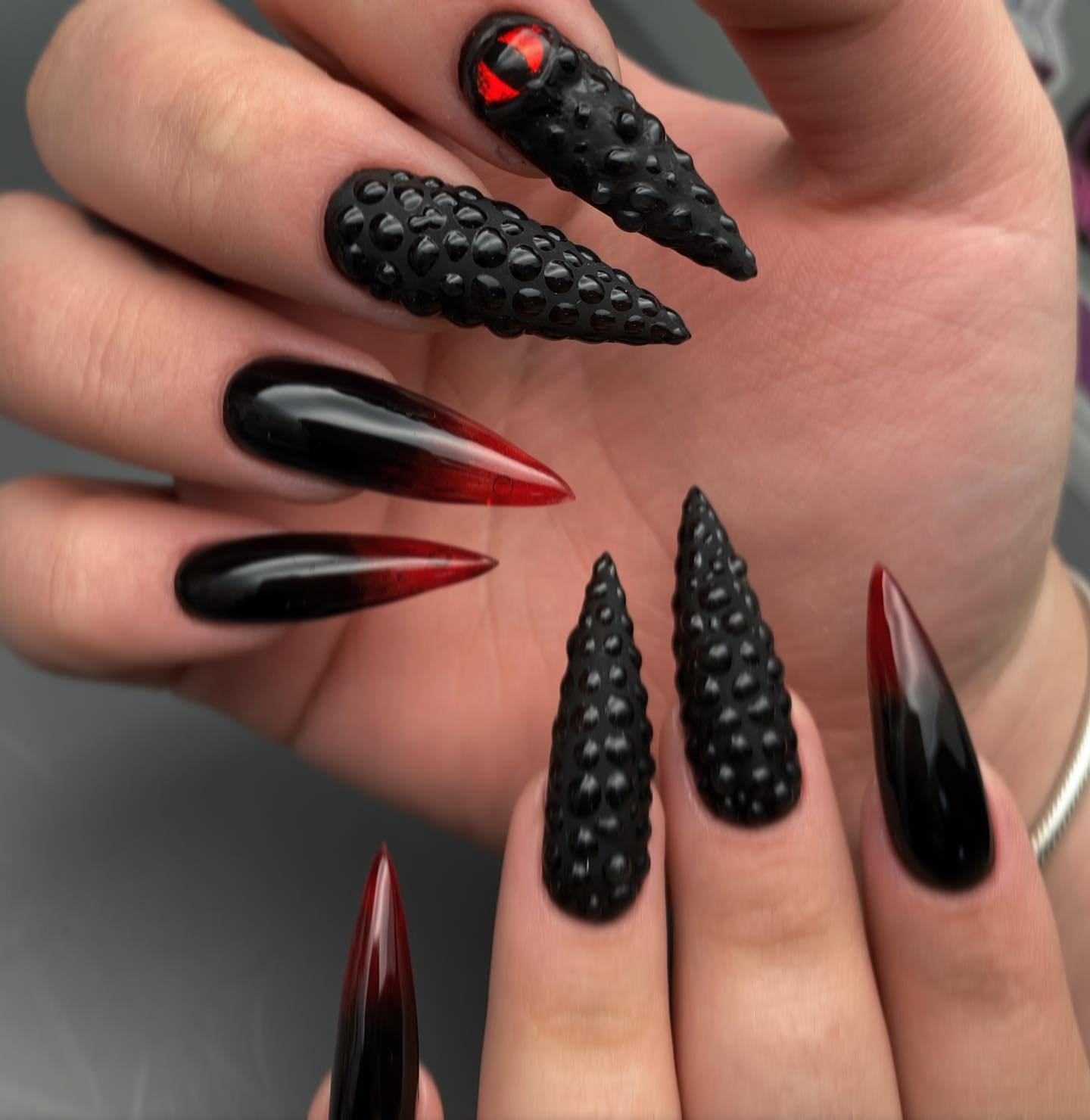 Long pointed nails mix smooth black polish with heavily textured black sections and sharp red accents.
