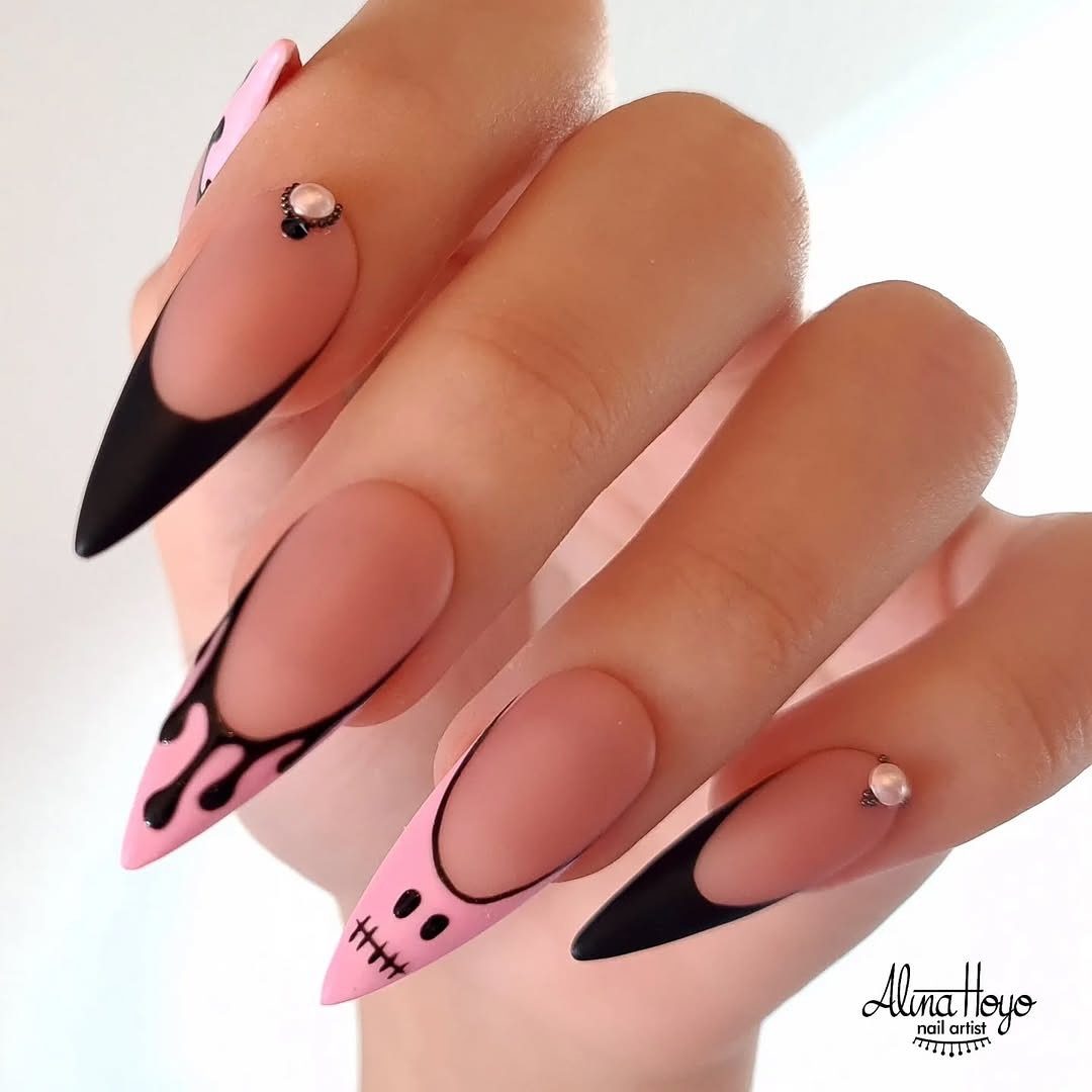 Pointed peachy nude nails feature black tips, small black-and-pink graphic accents, and tiny raised details.