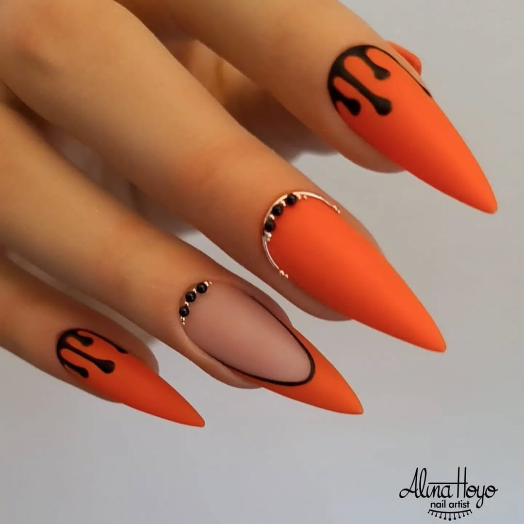 Long orange stiletto nails feature black graphic marks and small dark decorative accents.