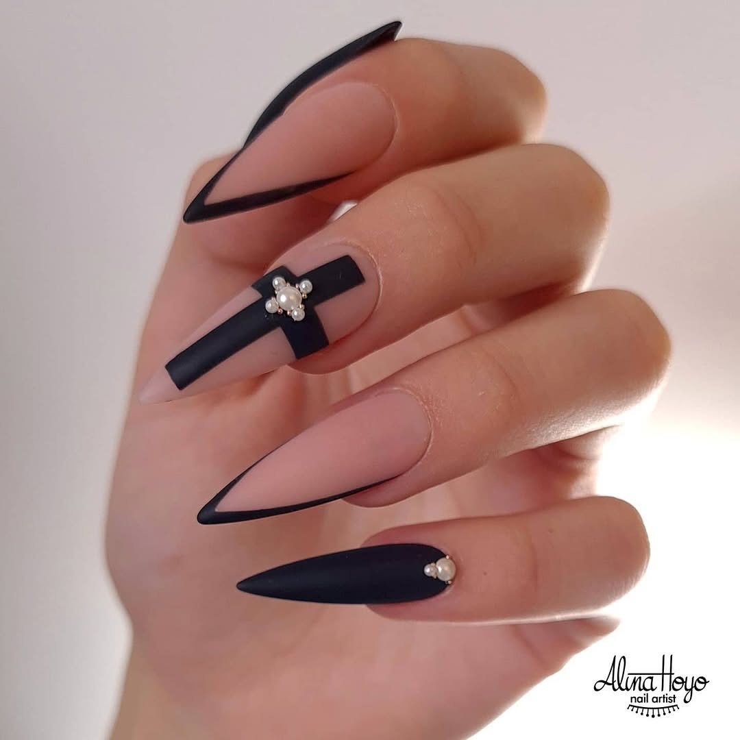 Pointed sheer pink nails feature fine black French outlines, a solid black tip, and a small cross-like charm.