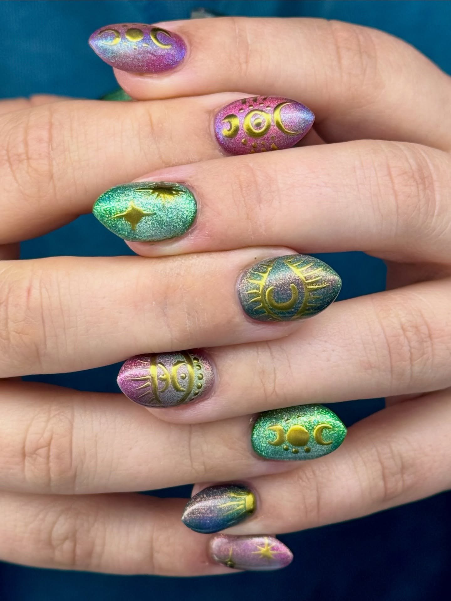 Short-to-medium oval nails show a multichrome purple-and-green shimmer with bright gold decorative accents on several nails.
